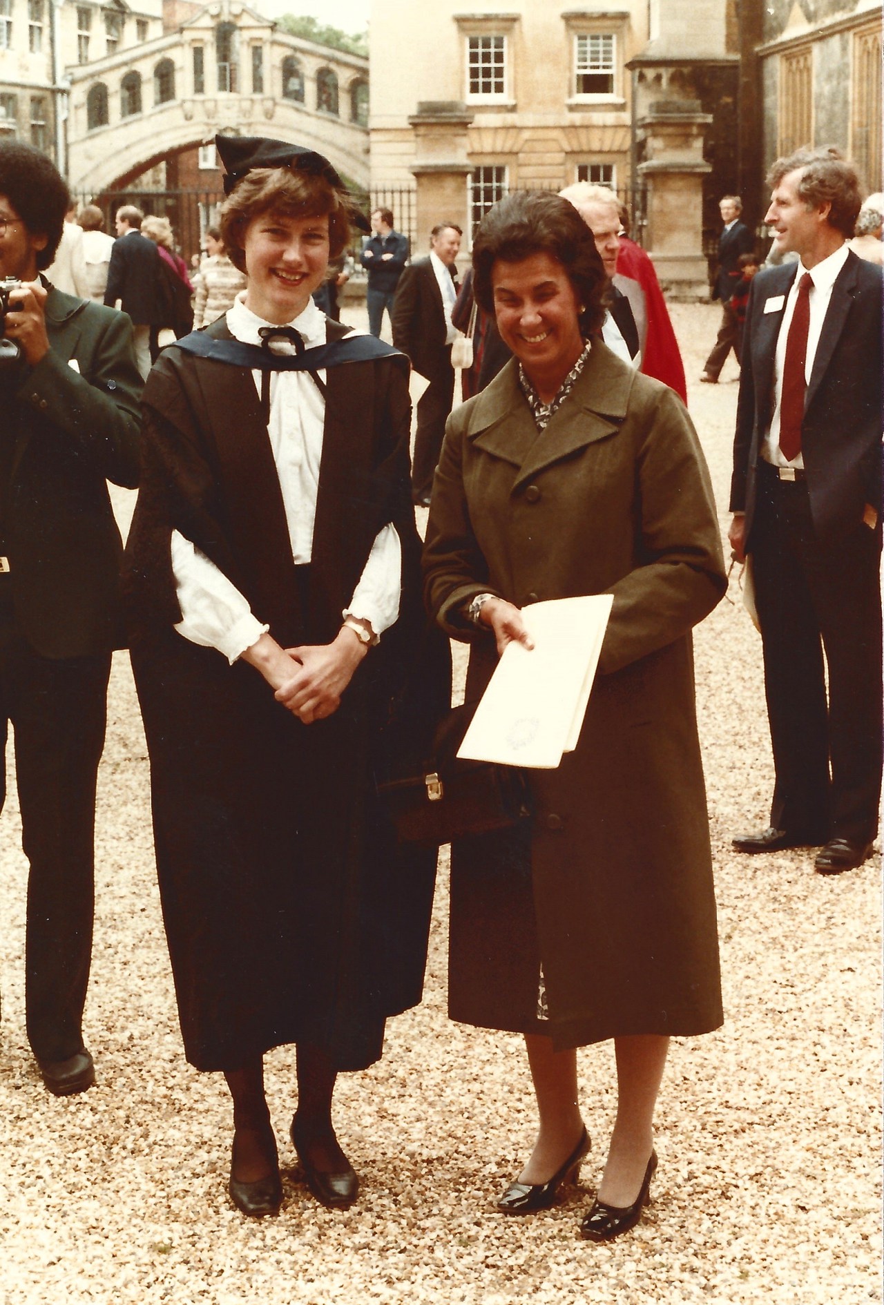 Margaret stood in graduation gown to the left of her mother, both smiling, with people walking behind them.