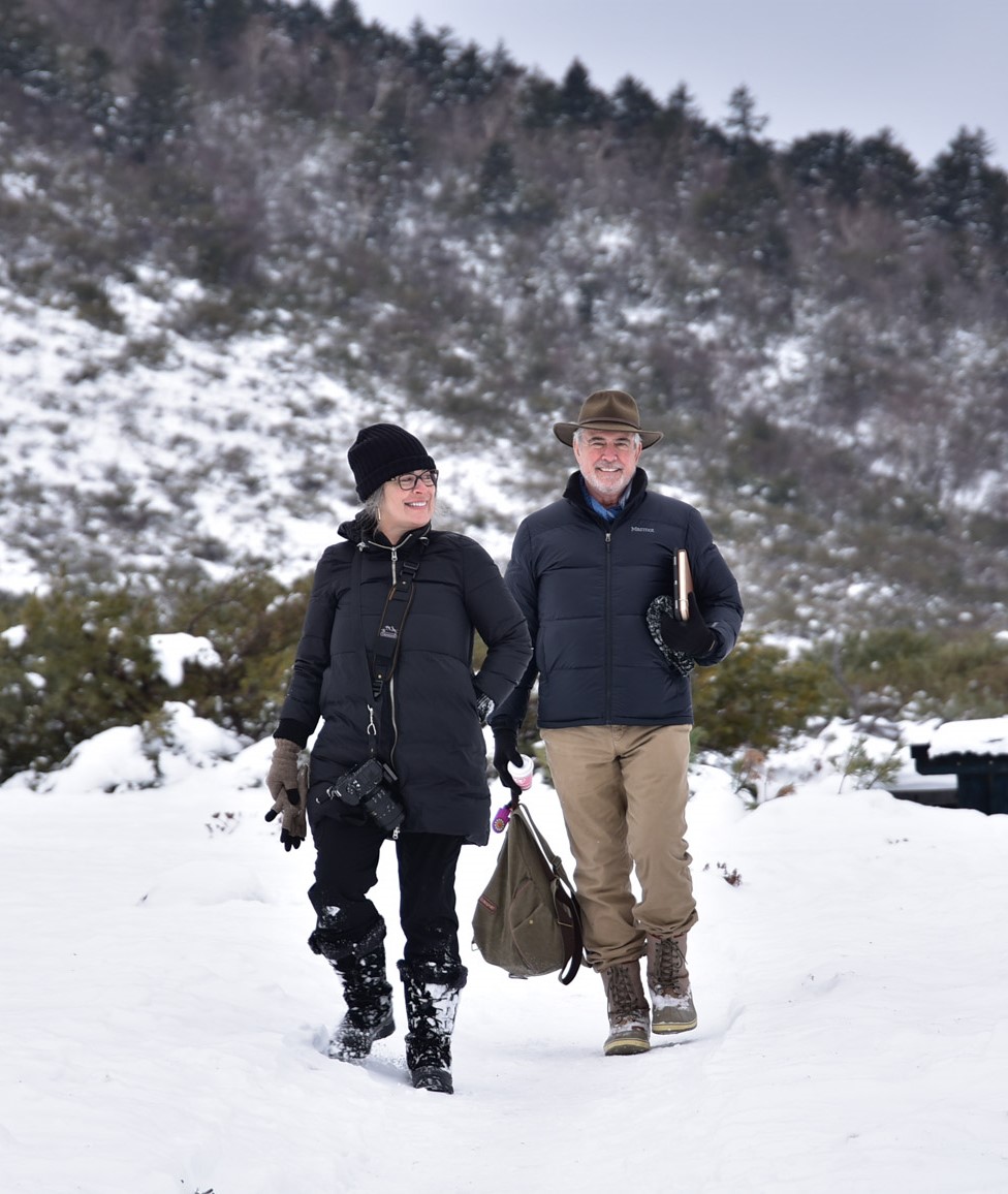 Elizabeth and Ken are pictured smiling as they walk throug a snowy field. A mountain full of snow covered trees is behind them.