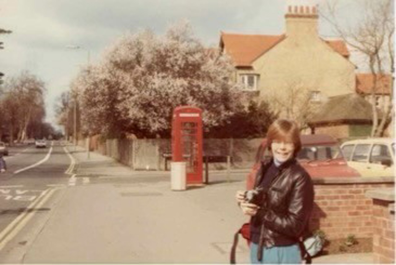 The picture shows Mary on a bright sunny day in Oxford. She is wearing a leather jacket. There are houses in the background, including a red telephone booth.
