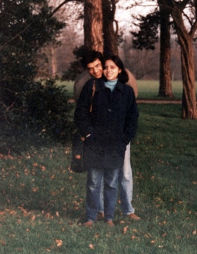 The picture shows Mukund and Soumya standing in the woods. Mukund is behind Soumya and his chin is resting on her shoulder. They are both smiling.