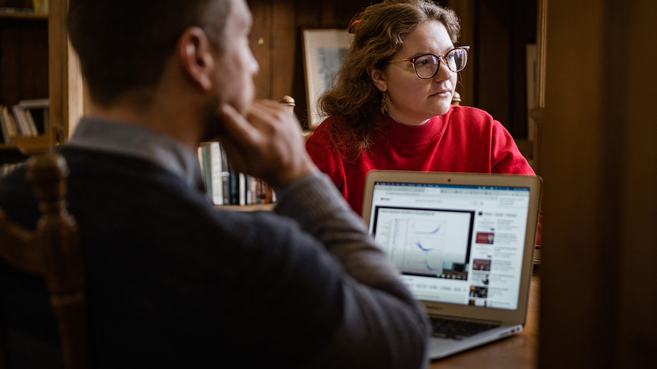 An image of two Rhodes scholars, a white man and woman, sitting opposite each other at a desk with an open laptop between them.