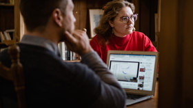 An image of two Rhodes scholars, a white man and woman, sitting opposite each other at a desk with an open laptop between them.