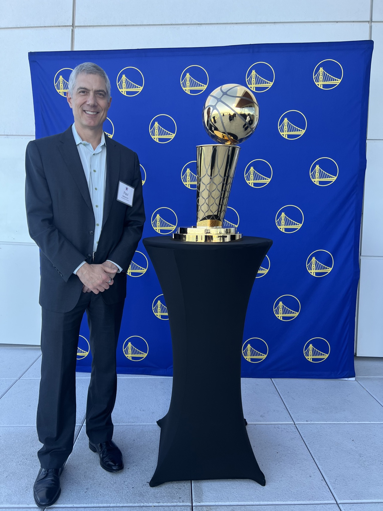 Paul stood in a suit posing next to a gold trophy which is placed on a black stand. The background is a blue display.