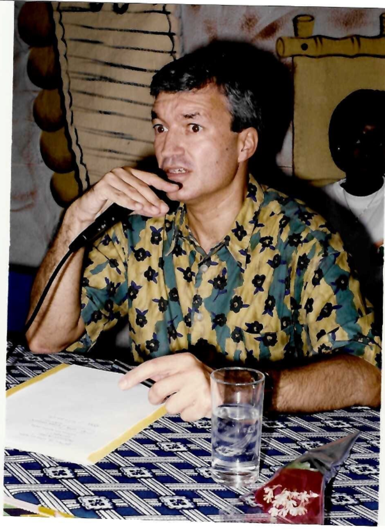 The picture shows Richard in a green-and-yellow shirt with dark patterns at a table with a microphone in his hand. In front of him is a file with papers.