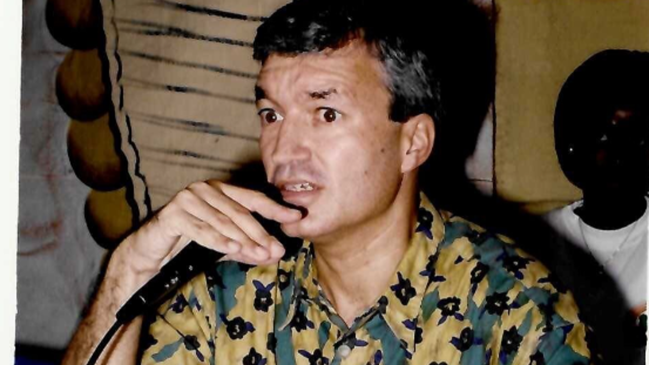 The picture shows Richard in a green-and-yellow shirt with dark patterns at a table with a microphone in his hand. In front of him is a file with papers.