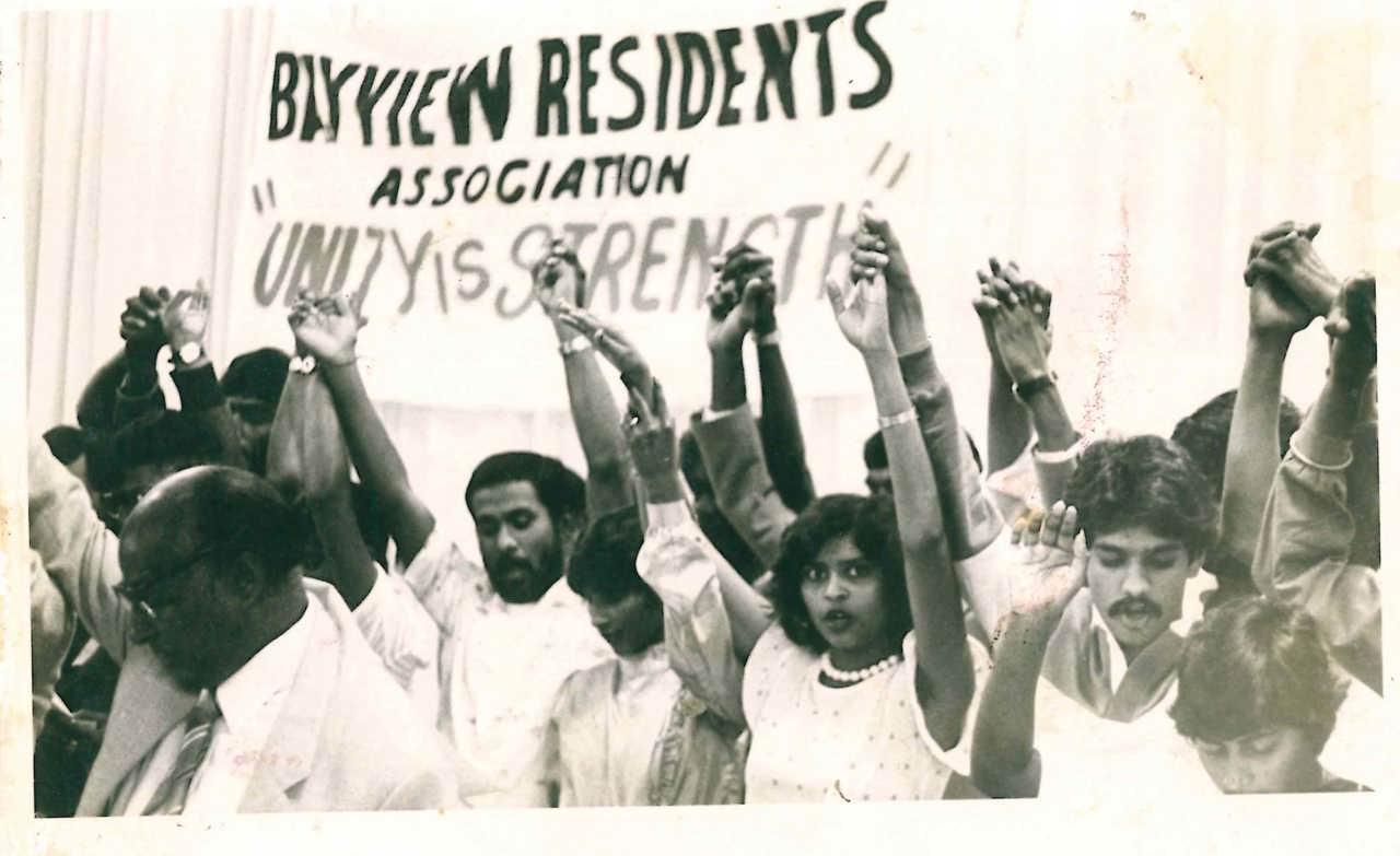 The picture shows Kumi protesting with other people. Their hands are in the air and they hold a banner on which is written 'Unity is Strength'