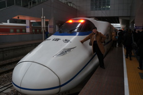 Elisabeth is seen leaning against a large, white bullet train at a train station.