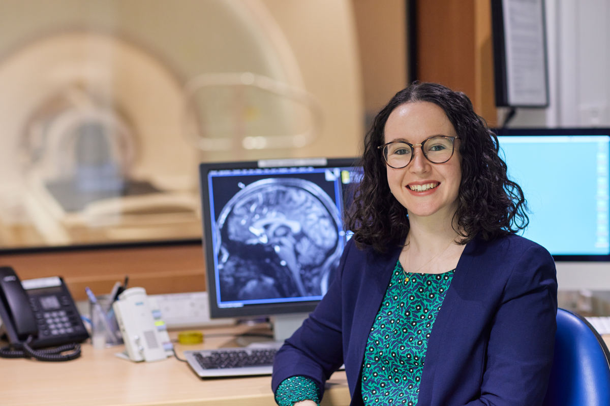 Image of a smiling woman in a blue jacket sitting in front of a computer screen that shows an x-ray scan of a brain.