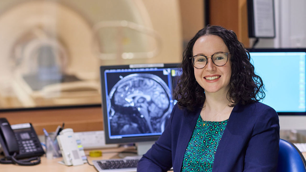 Image of a smiling woman in a blue jacket sitting in front of a computer screen that shows an x-ray scan of a brain.