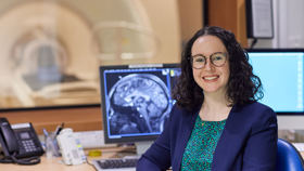 Image of a smiling woman in a blue jacket sitting in front of a computer screen that shows an x-ray scan of a brain.