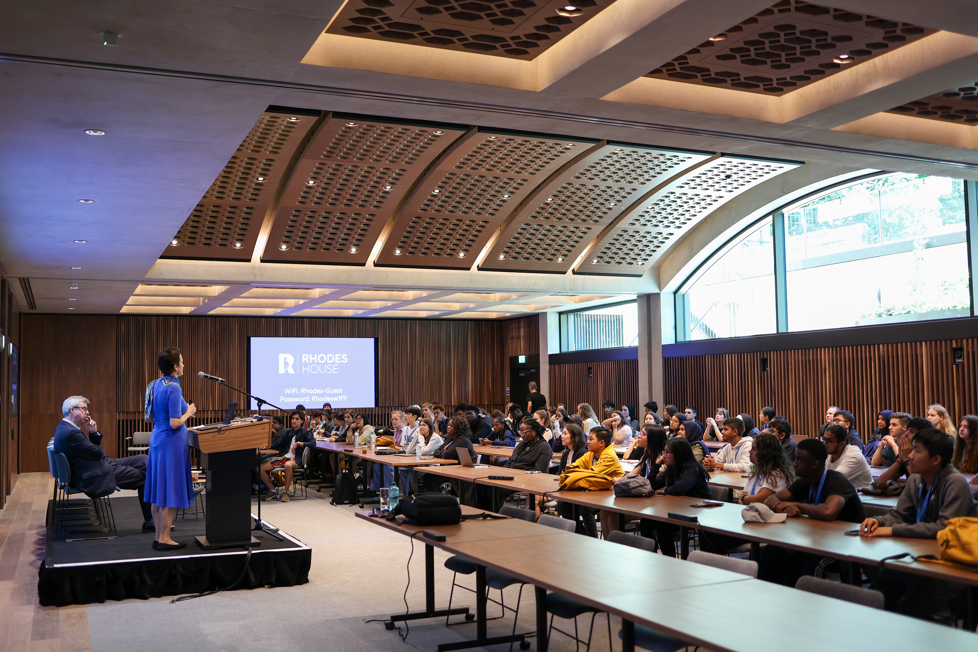 An image of Dr Elizabeth Kiss, the warden of Rhodes House, making a speech at a podium to a conference room of students seated at desks.