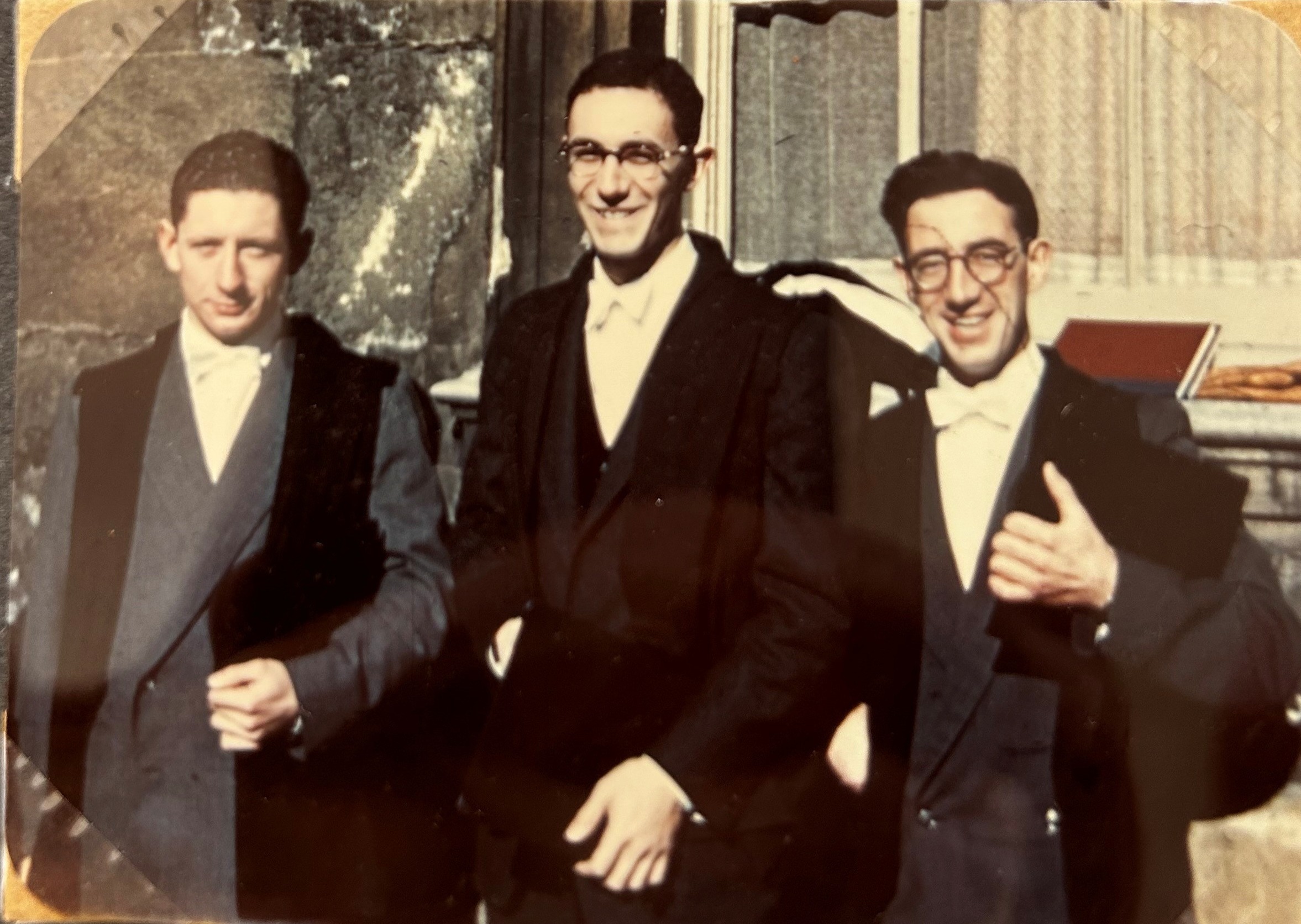 3 young men stand smiling side my side in matriculation suits and gown. All wear a white bow tie.