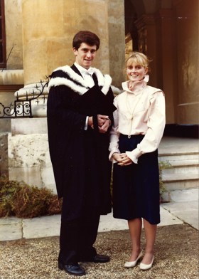 A picture of John in a graduation gown standing next to his wife who is wearing a shirt and a skirt.