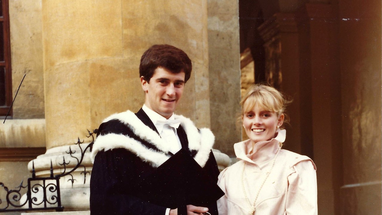 A picture of John in a graduation gown standing next to his wife who is wearing a shirt and a skirt.
