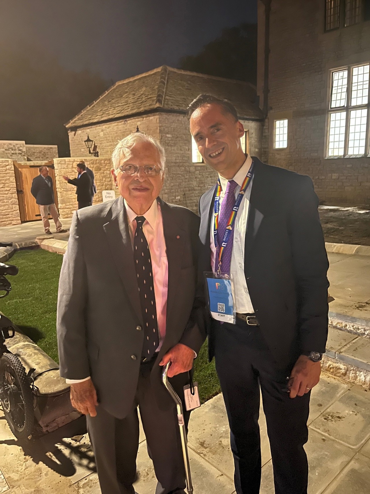 A picture showing John in a suit standing next to Rodolfo, also in a suit, with the Rhodes House in background