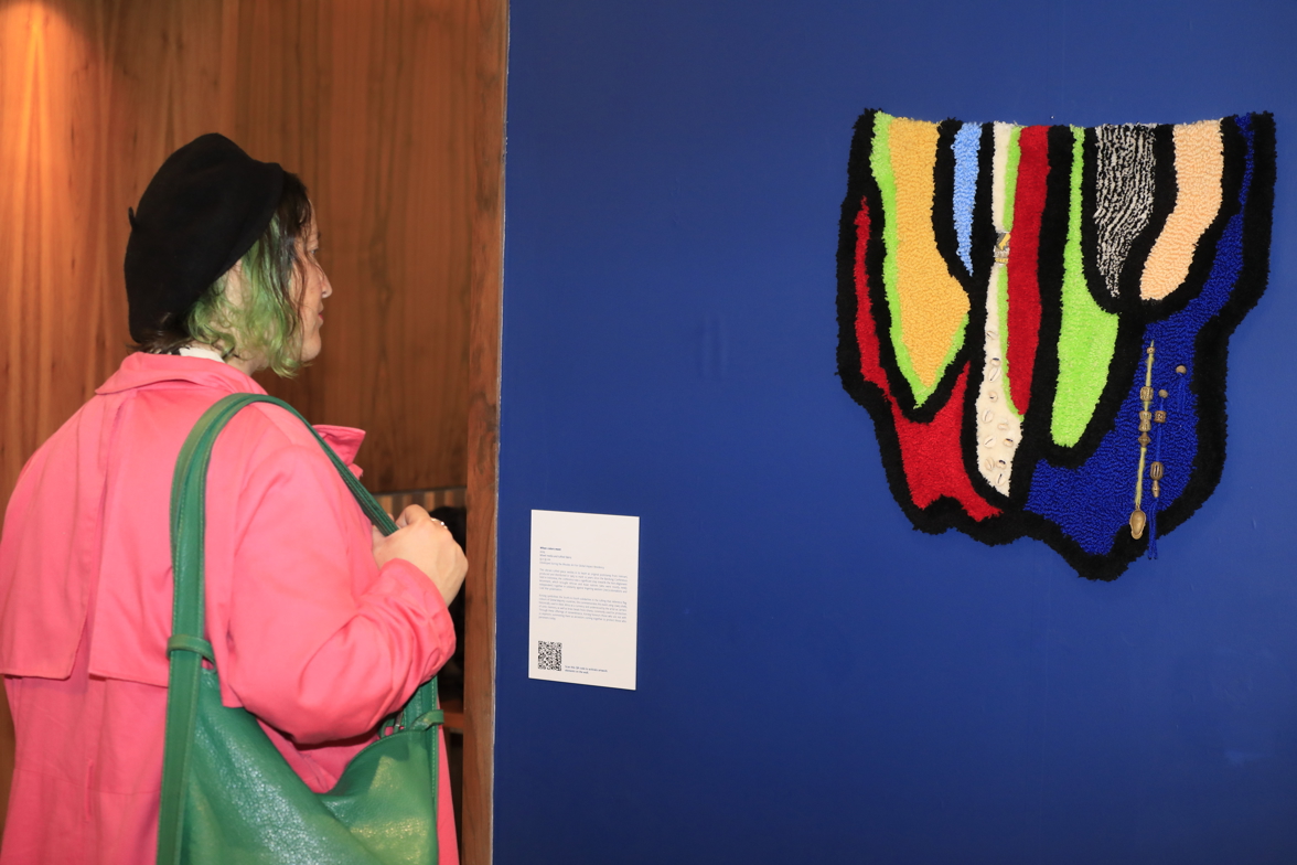 A visitor wearing a pink jacket looks at a colourful tufted fabric artwork on a blue wall.