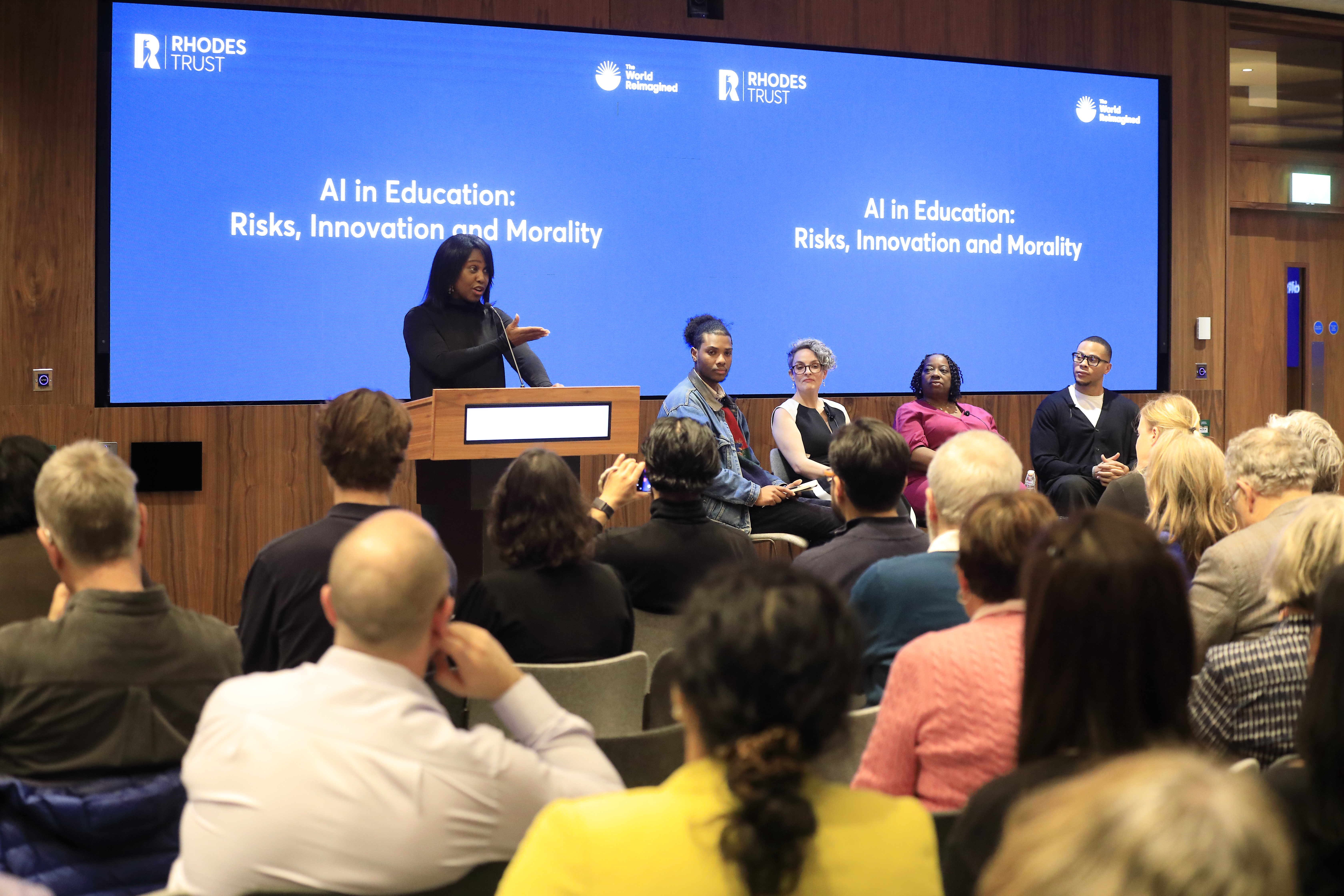 A speaker at a lectern in front of 4 speakers and a blue screen
