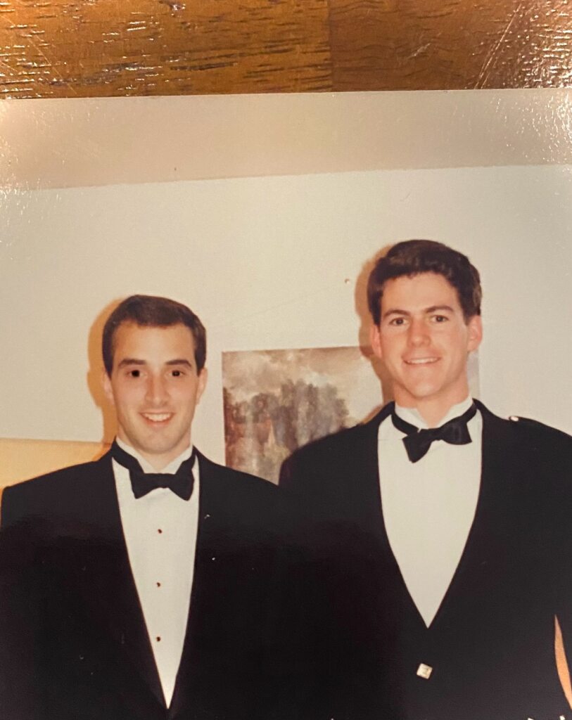 David and Tom both stood in a suit and bow tie, smiling. The photo is taken from their chest upwards, and there is a framed image behind them, although what is pictured is not clear.