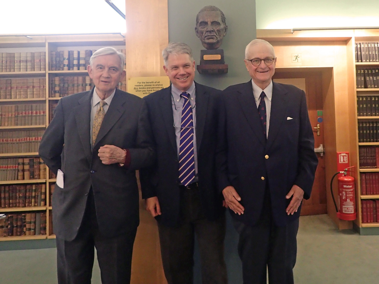 Three men stood in a library, wearing suits and smiling into camera.
