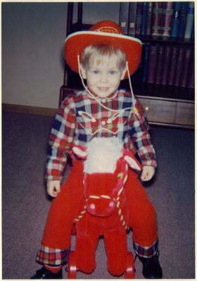 Eric as a toddler wearing a red cowboy hat, red checked shirt, red trousers, sitting on a red play horse and smiling.