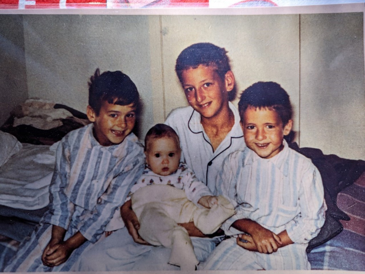 A picture showing three little boys in their pajamas holding an infant. They are all smiling at the camera.