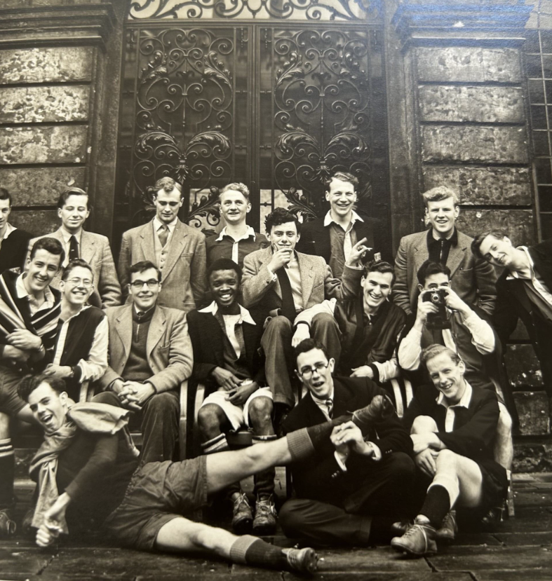 Black and white photo of a football team posing. All are smiling, some standing, sitting, and one young man lies across the floor in front of them