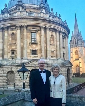 The picture shows Ralph in a black suit and Betsy in a white top and black pants, standing in front of the Radcliffe Camera. They are both smiling.