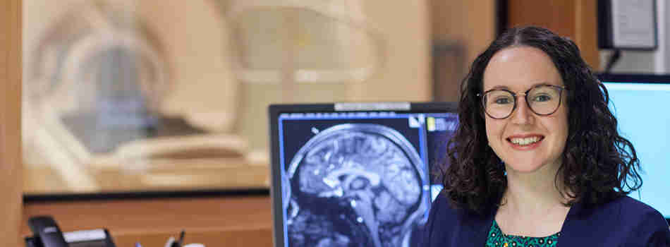 A woman smiling at the camera against the backdrop of a medical scanner with a brain scan on a screen behind