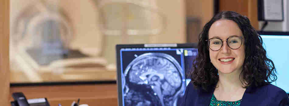 A woman smiling at the camera against the backdrop of a medical scanner with a brain scan on a screen behind