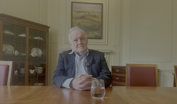Gavin is pictured sat in a suit at a table. There is a glass of water in front of him, and behind him is a watercolour painitng, and a cabinet displaying china.