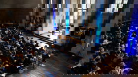 McCall MacBain Hall conference with delegates facing the speaker in the bay window, with screens on either side.