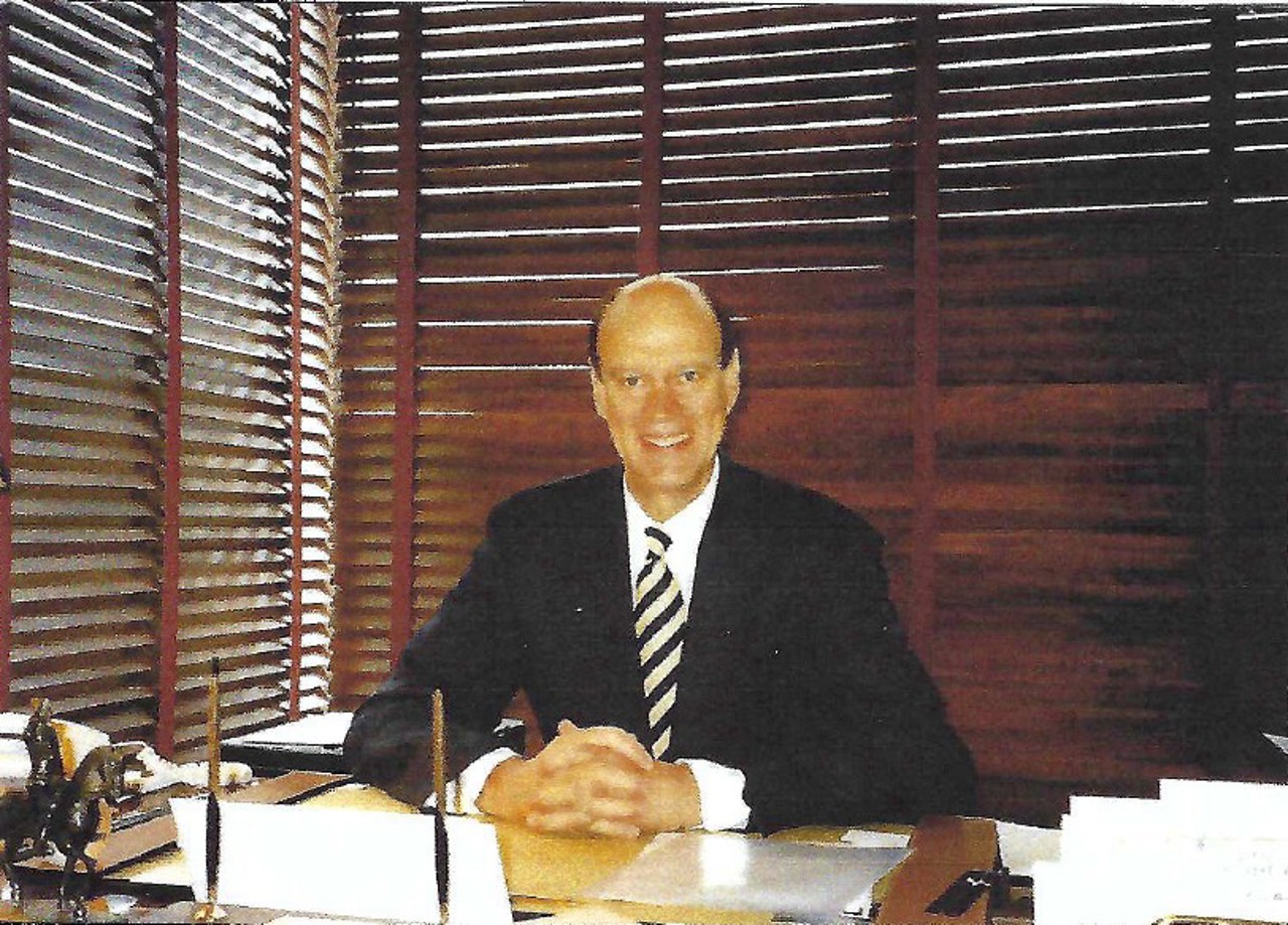 Willie is sat in a suit at a desk, hands together and smiling. The windows are covered by brown shutters behind him.