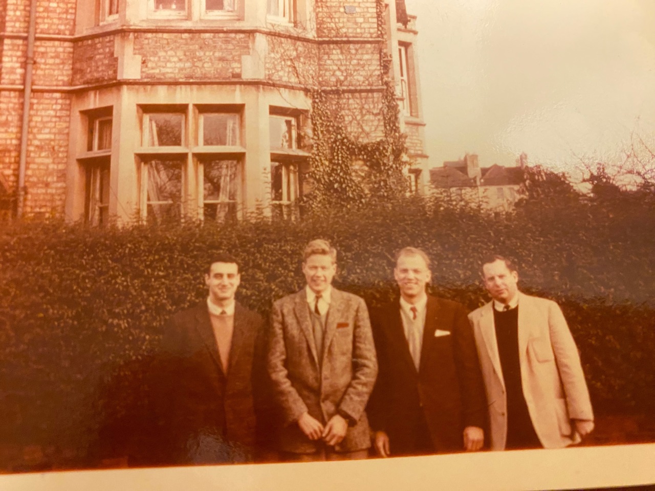 Four young male students stood in blazers before a house and a bush.