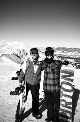 Black and white photo of father and son holding snowboards on top of a mountain.