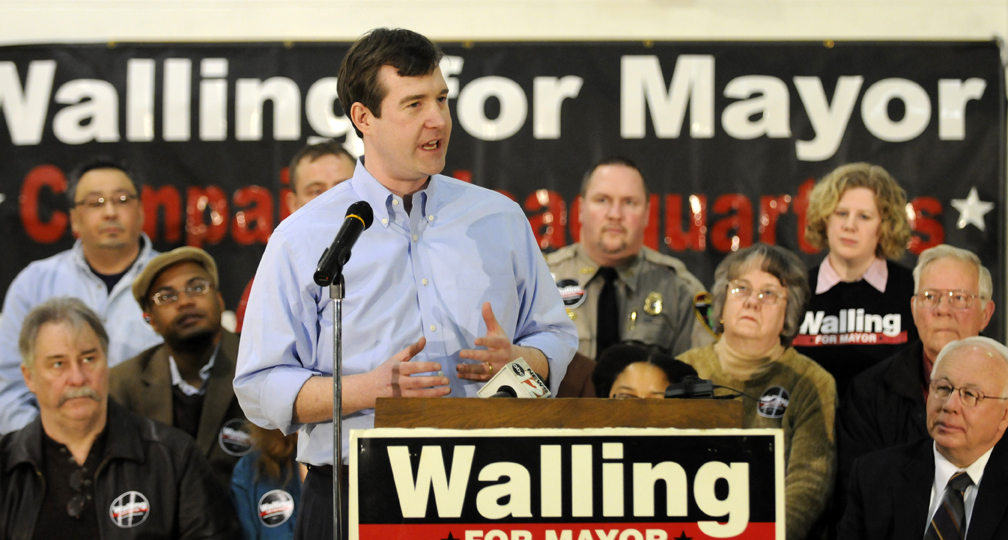 Dayne stands before a lecturn which reads 'Walling For Mayer'. The poster behind him says the same. He is talking to a crowd through a microphone. There is a group of people sat behind him watching him speak.