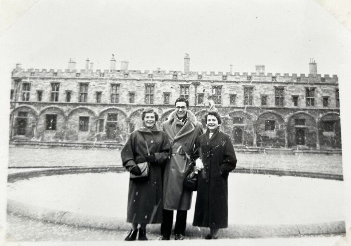 Black and white photo of Ken stood in a large fur coat between two ladies. They are before the christ church fountain in the centre of the college quad