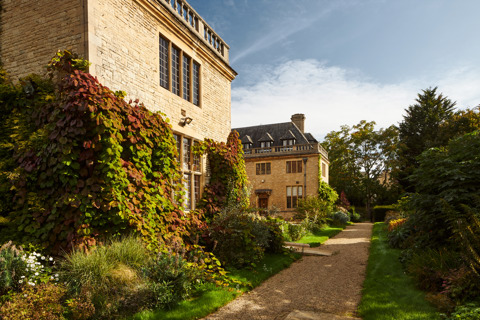 An image of the exterior of Rhodes House on a sunny day. The stone walls are covered in ivy and climbing plants, and there is a gravel path through a garden lined with hedges.