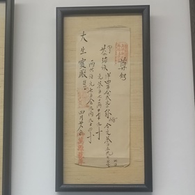 Photo is of a framed piece of paper, which appears to be very ancient and has handdrawn black Chinese characters with a few red prints.