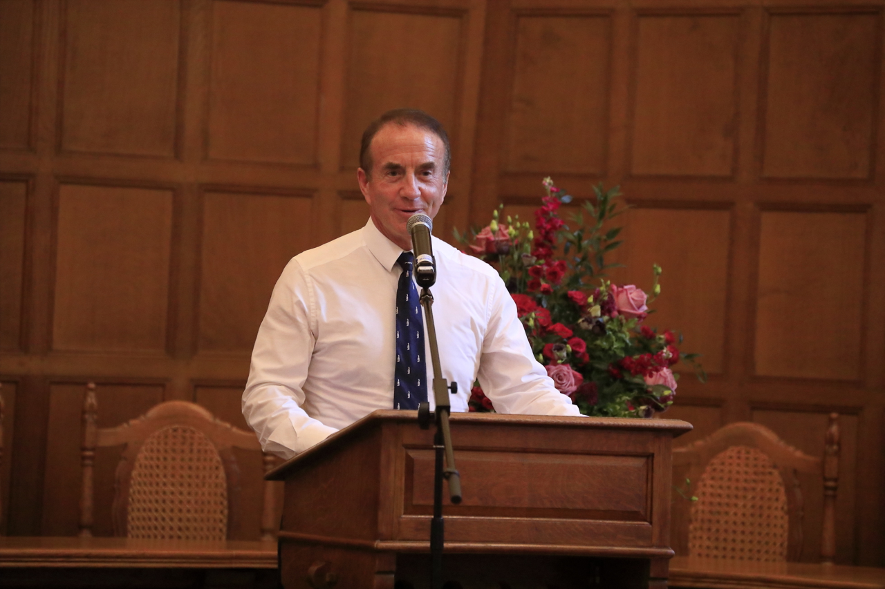 John stood in a white shirt and tie before a podium and mic. There are pink flowers in a vase behind him.