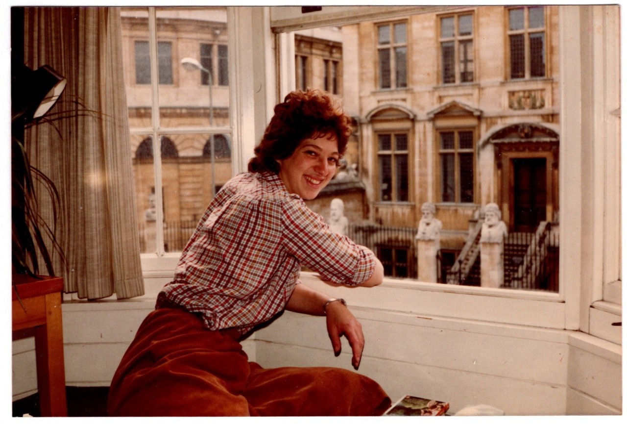 The picture shows a young Joy leaning against a window with sandstone buildings in the view outside