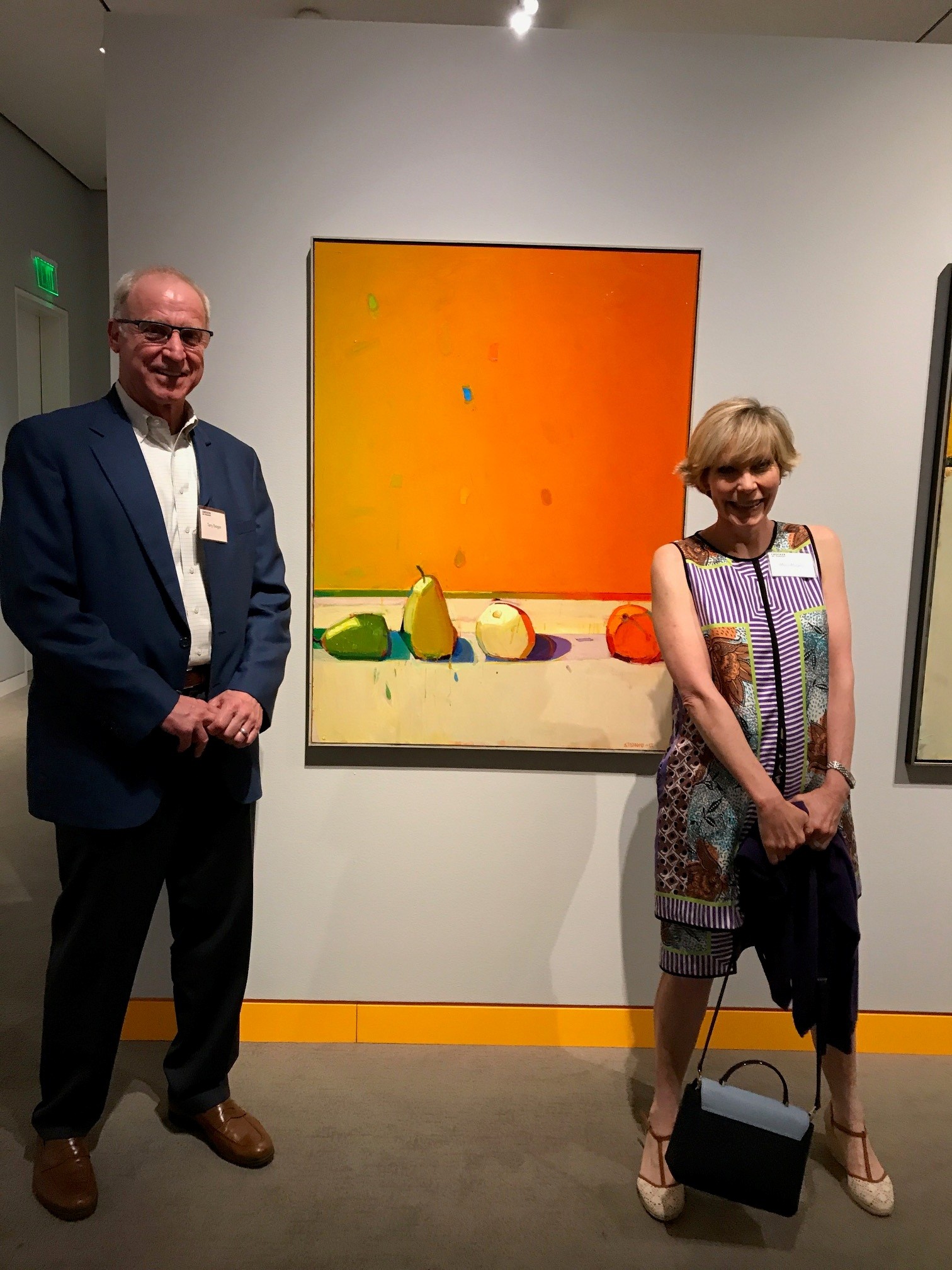 Mary stood in a dress to the right of an orange painting with four fruits on it (a pear, orange, apple, and unclear fruit). To the left of the painting stands her husband wearing a suit.