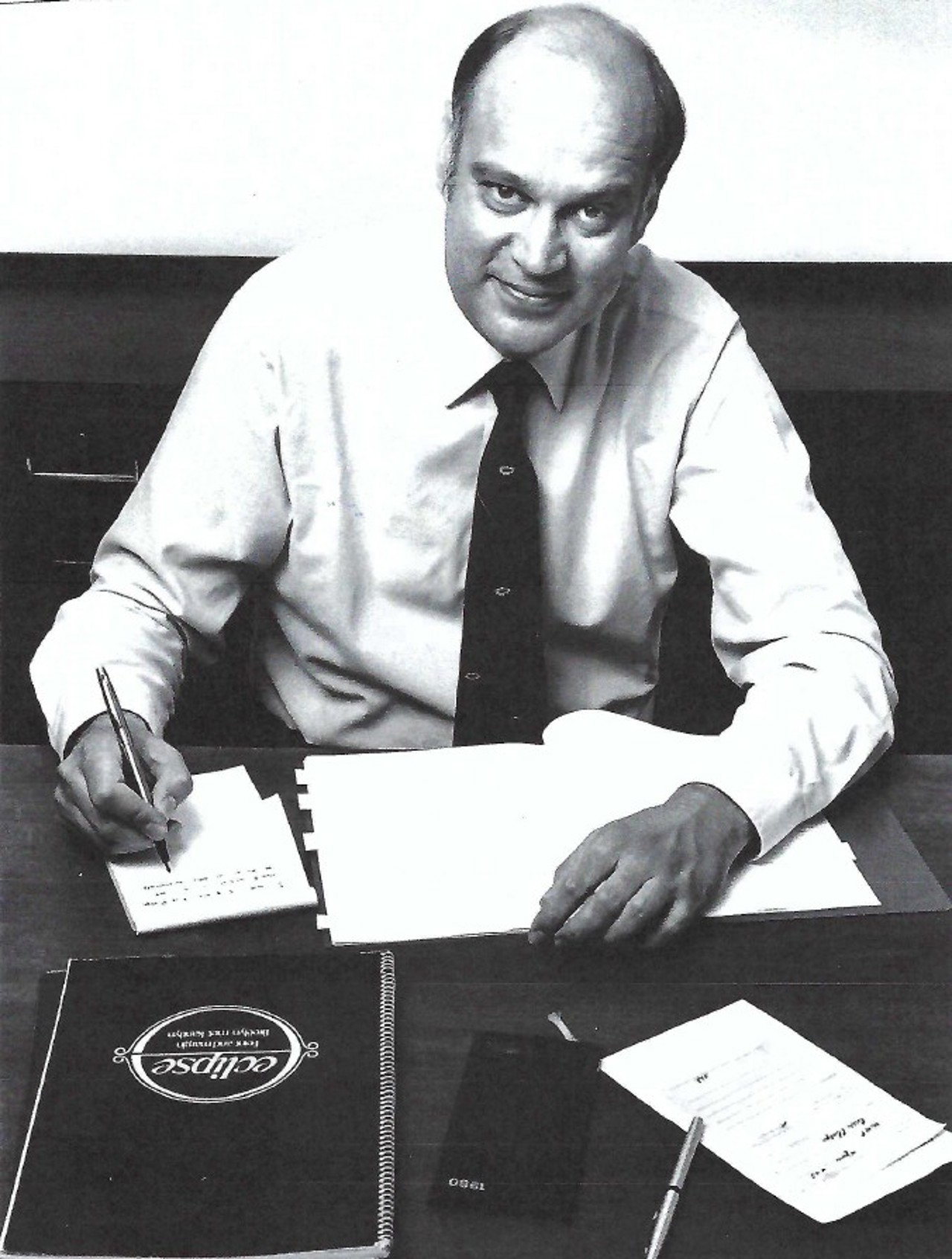 Black and white photo of Willie in a white shirt and tie, sat at a desk and writing. He is looking up to the camera whilst holding a pen.