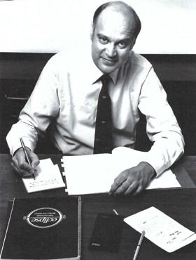 Black and white photo of Willie in a white shirt and tie, sat at a desk and writing. He is looking up to the camera whilst holding a pen.