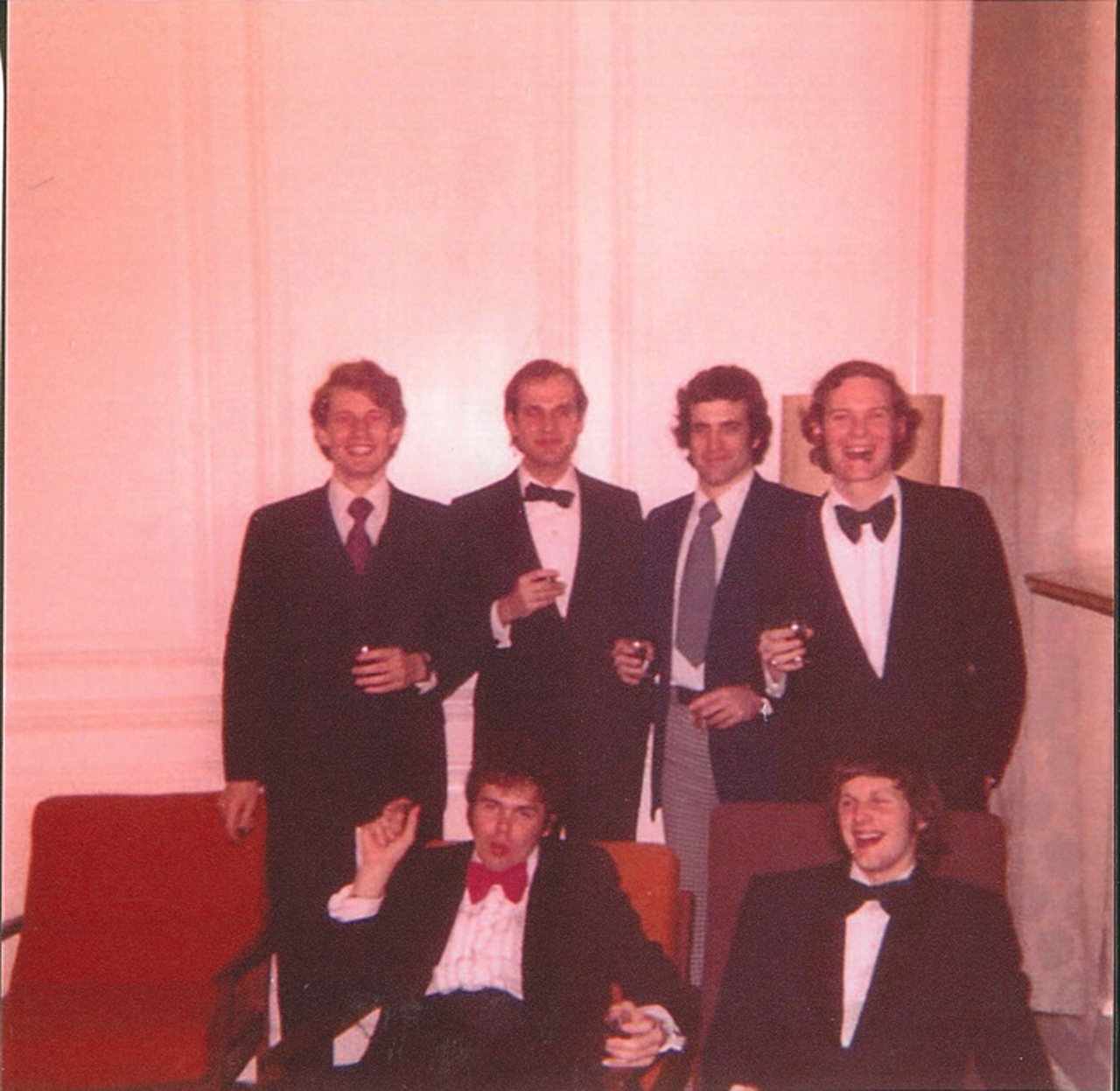 A picture showing six people in formal attire. They are all wearing bowties. Three of them are standing and two are sitting on a red couch.