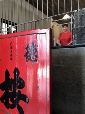 Elisabeth is seen standing on a platform int he distance. There are bars in front of her which appear to replicate an ancient pawnshop. There is a large print in front of her which is bright red and has Mandarin characters painted on it.