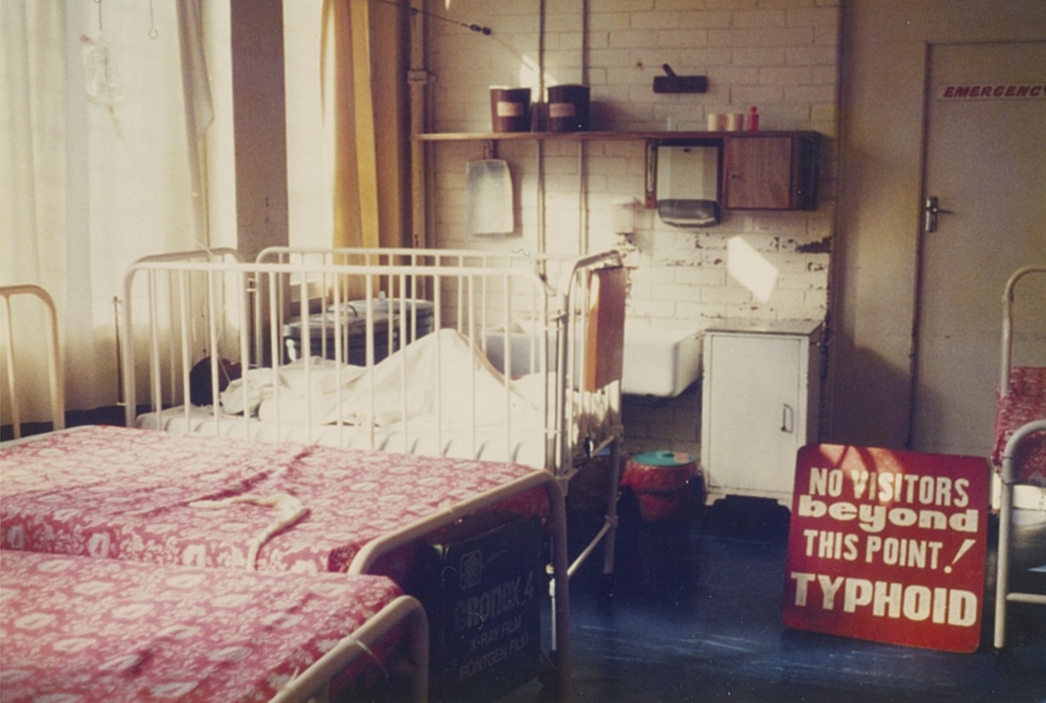 Photograph of a typhoid ward. Children's beds are seen, and a cot in the back with a child lying in it. The sign on the right reads 'No visitors beyond this point! Typhoid'