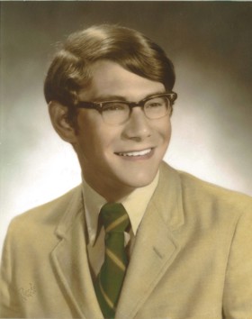 Formal school photo of younger Peter wearing a beige suit, tartan tie, and smiling.