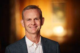 Headshot of Mark, in a white shirt and grey coat. He is smiling.