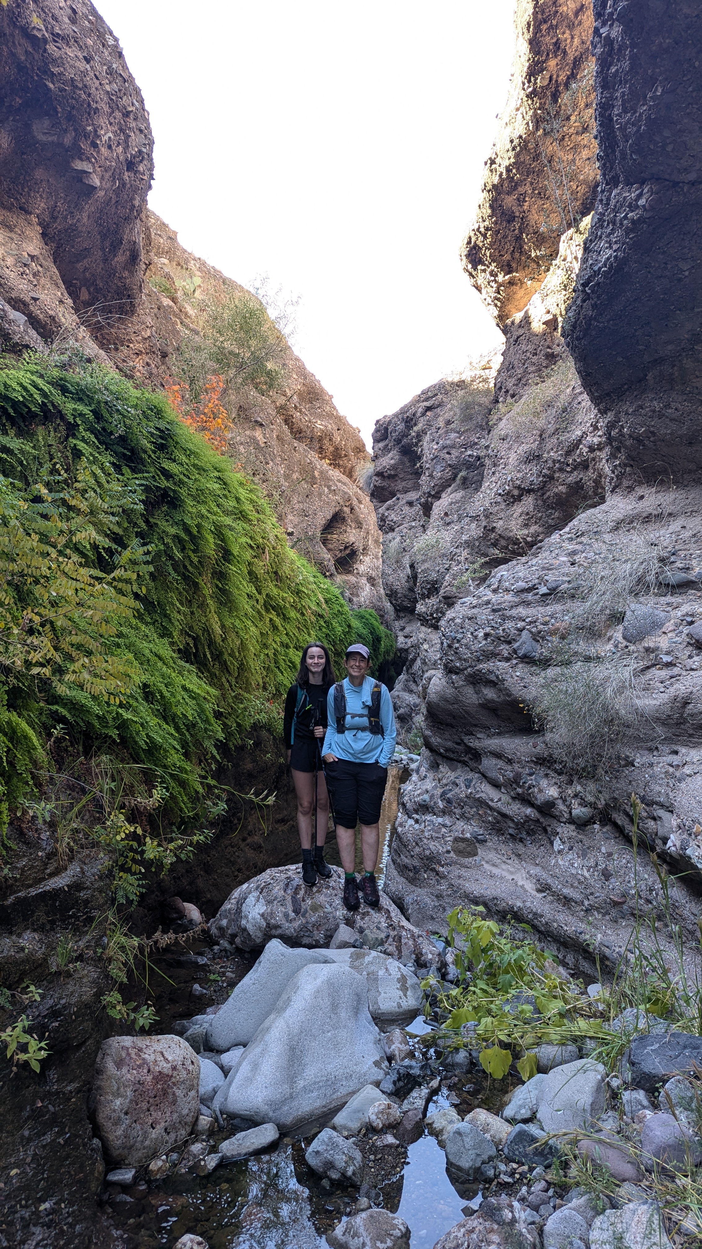 Jane stood to the right of a young girl, they are in between large boulders and stood on rocks. Both are wearing backpacks.
