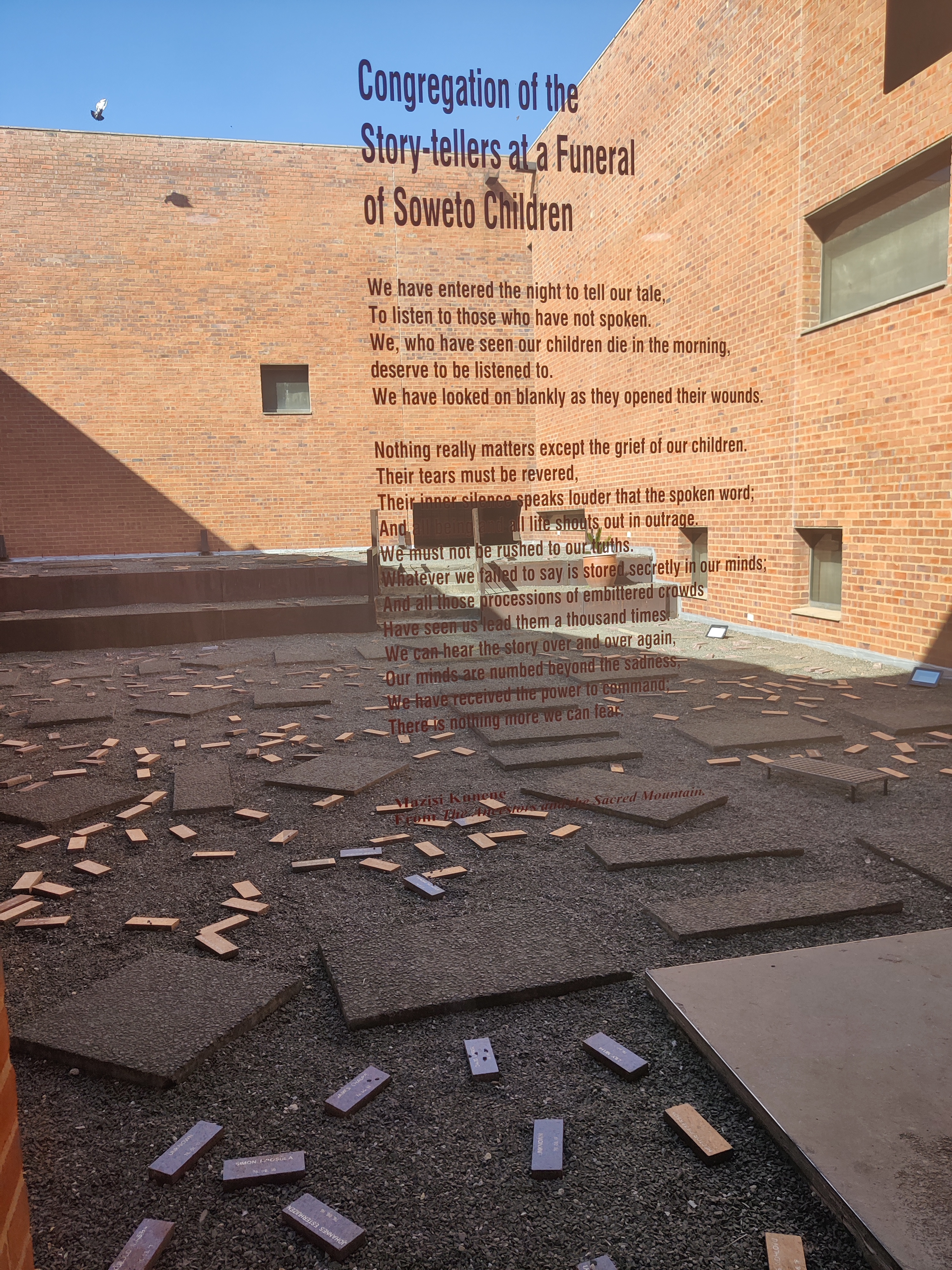 At the Hector Pieterson Museum in Soweto, behind a glass wall bearing the title and text of Mazisi Kunene’s poem ‘Congregation of the Story-tellers at a Funeral of Soweto Children’, lie bricks with names amongst larger grave like shapes on the ground.